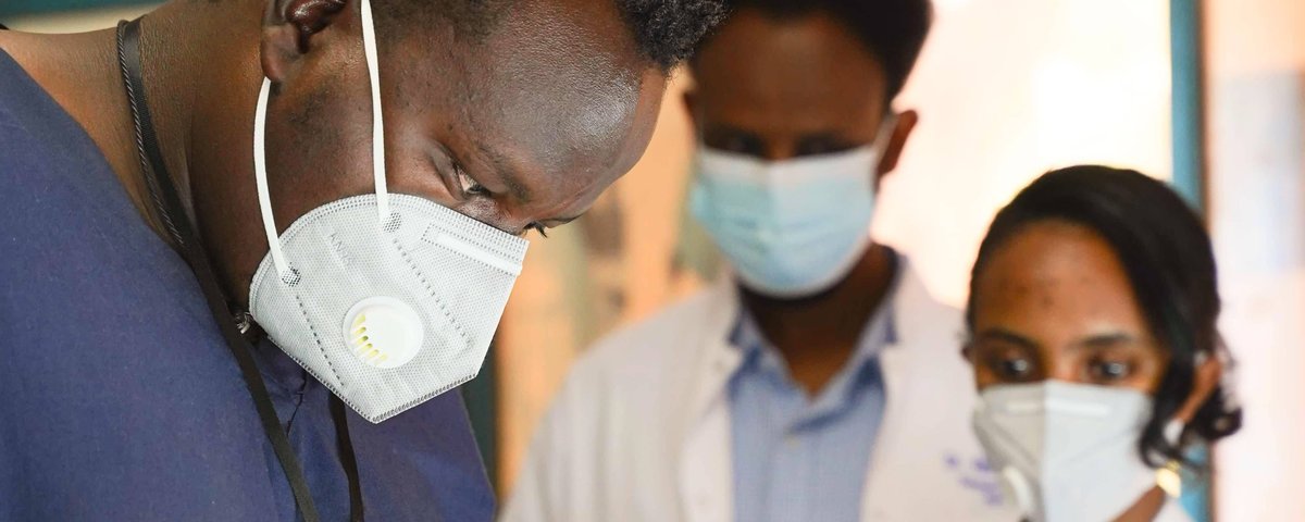 A Doctor wearing a mask, working on something out of shot, with two other doctors in the background watching him work
