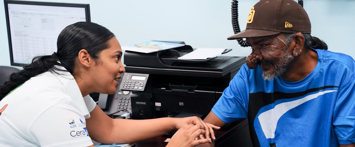 A man with a skin condition across his nose is being treated by a clinician from the ILDS, provided by GLODERM funding. The clinician is treating the man with a friendly warm smile.