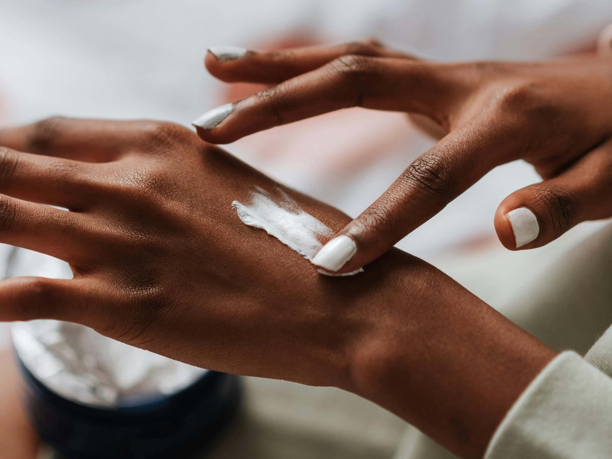 A woman with dark skin is applying cream to the back of her hand with her pointer finger.