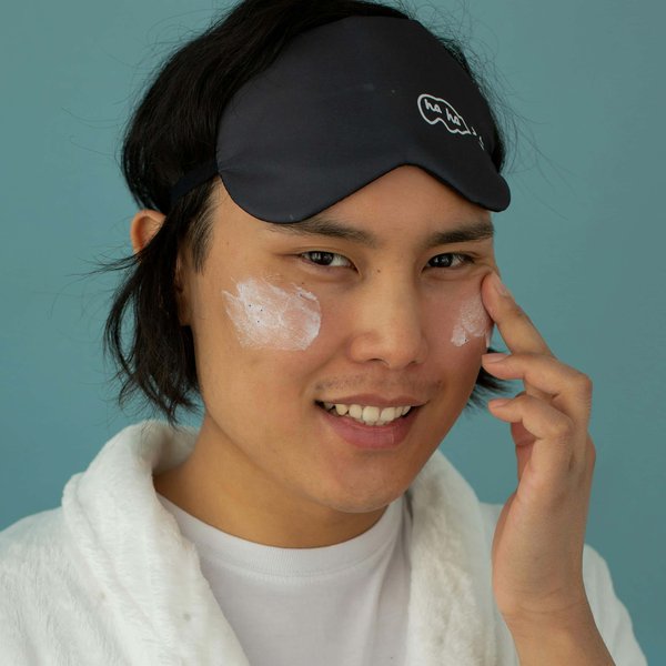 A man is wearing an eyemask above his forehead and applying cream to his cheeks